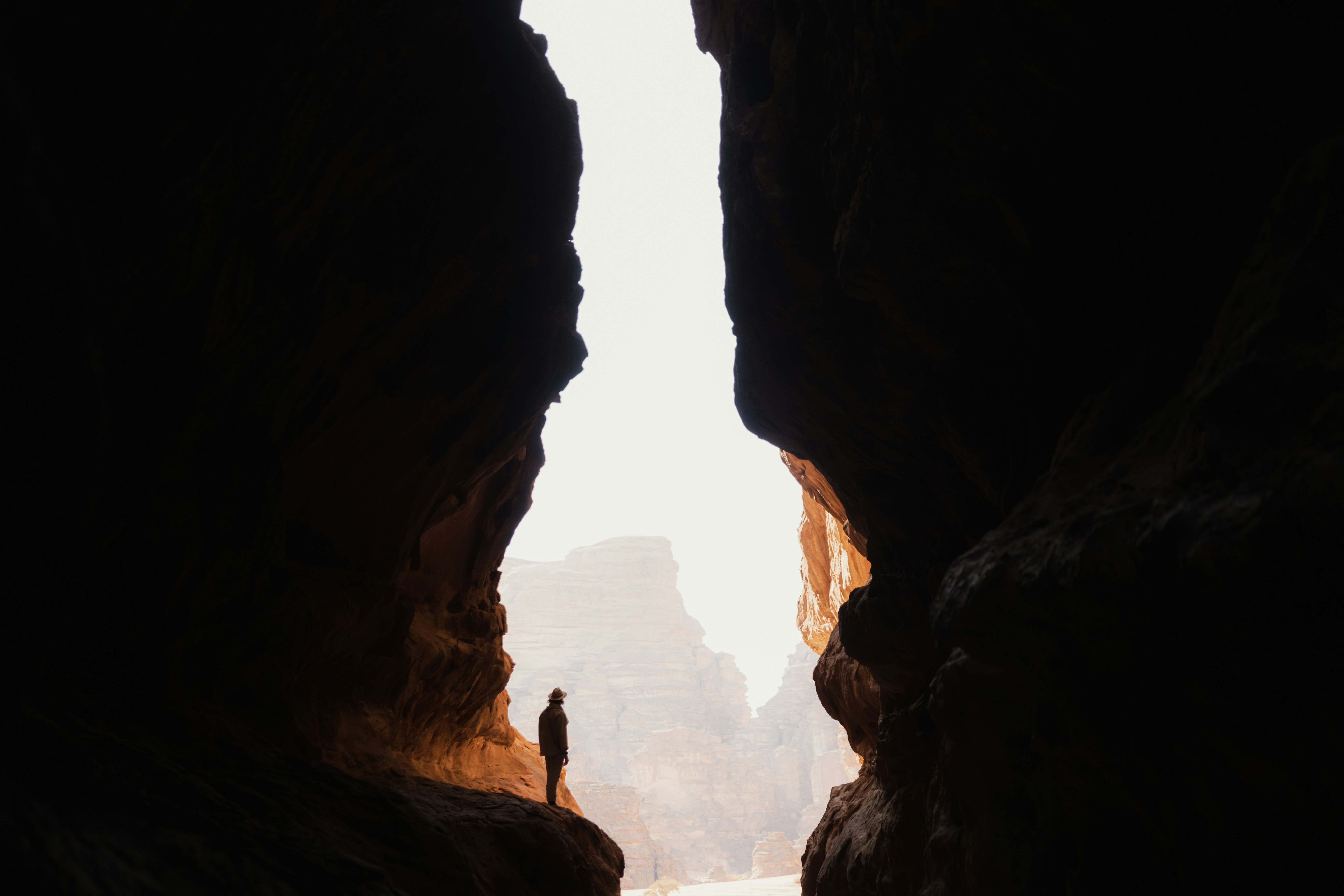 A person silhouetted in a dark cave opening, looking out toward a bright sunlit canyon