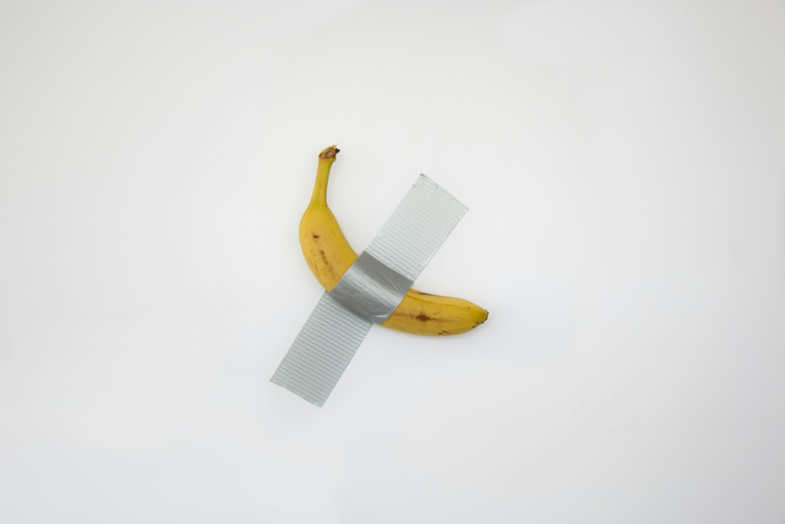 A yellow banana taped to a white wall with a single strip of grey duct tape