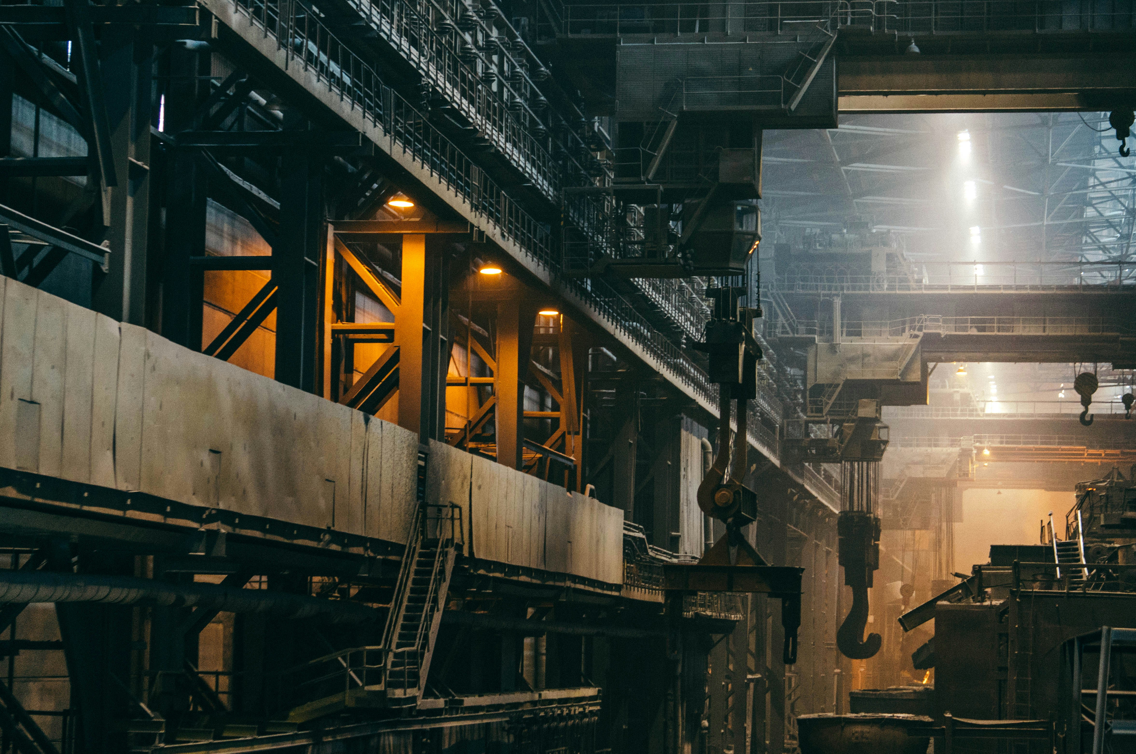 The dim interior of a large industrial factory with heavy machinery, gantries, and a crane hook