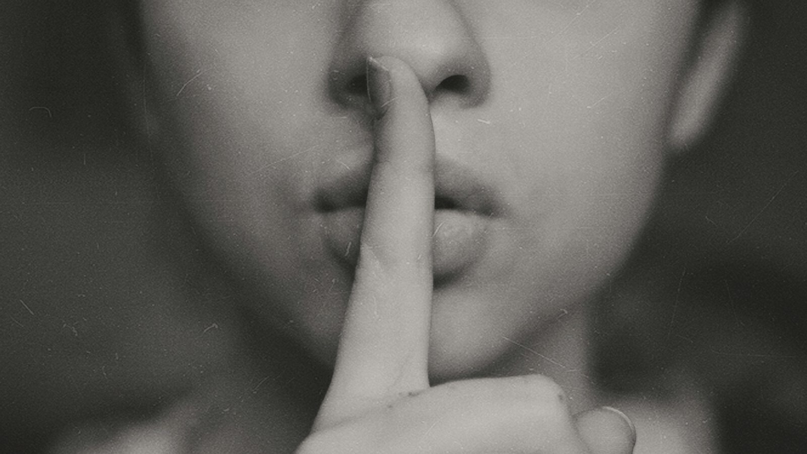 A grayscale photo of a woman with a finger to her lips, signaling silence