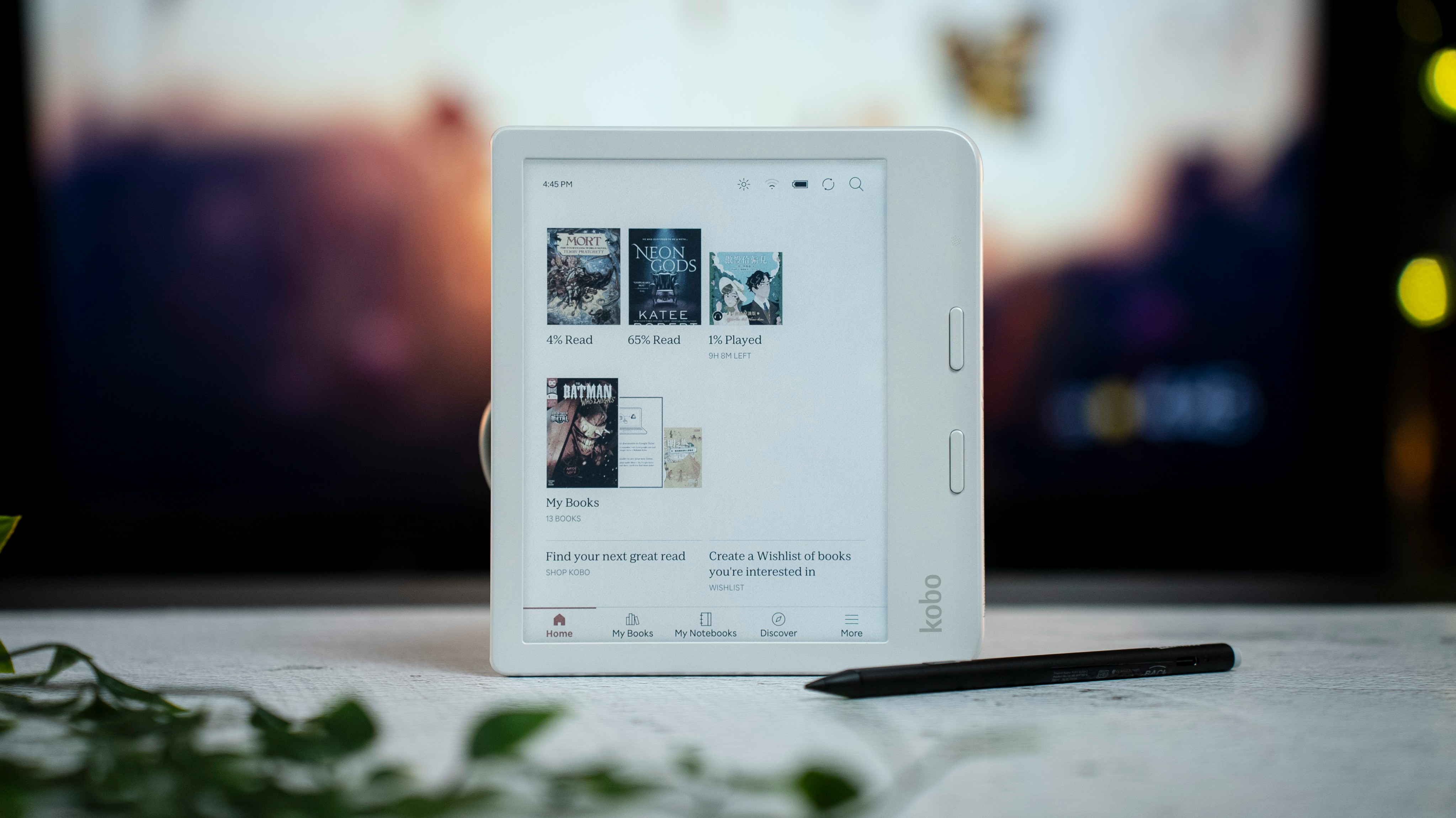 A white Kobo e-reader on a table showing its home screen of books, a stylus beside it and a plant in the foreground