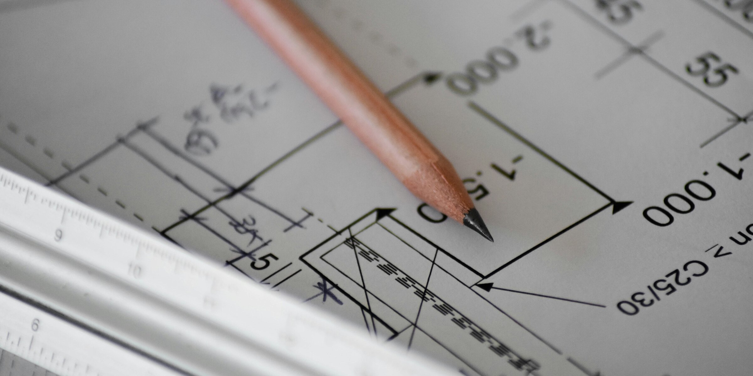 A sharpened wooden pencil resting on a printed technical drawing with dimension lines and measurements, a white scale ruler across the lower left corner