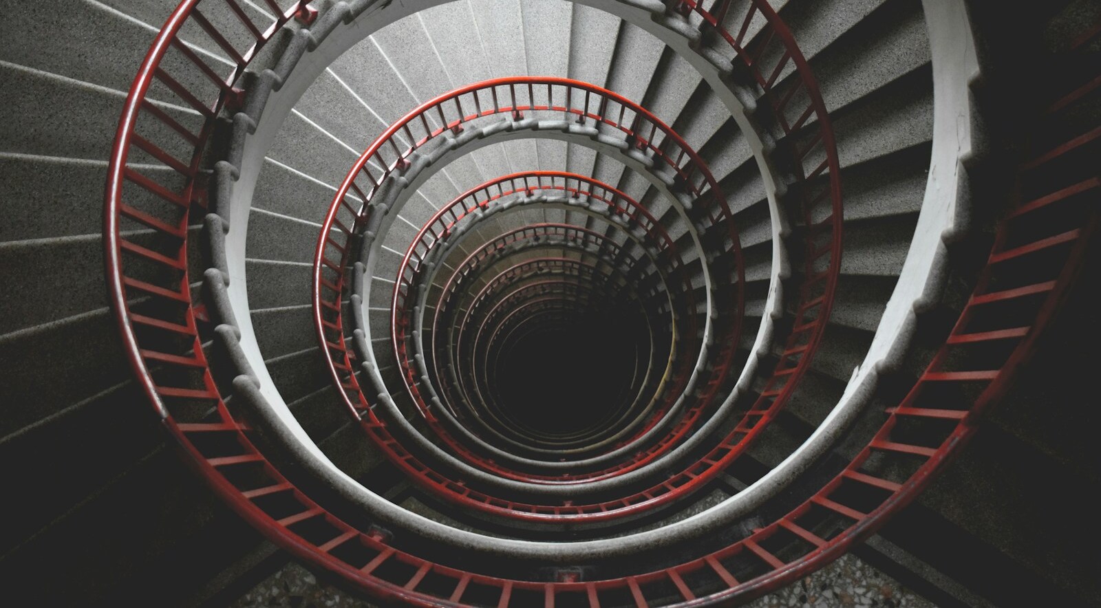 A spiral concrete staircase with red railings viewed from above, looking down through nested loops