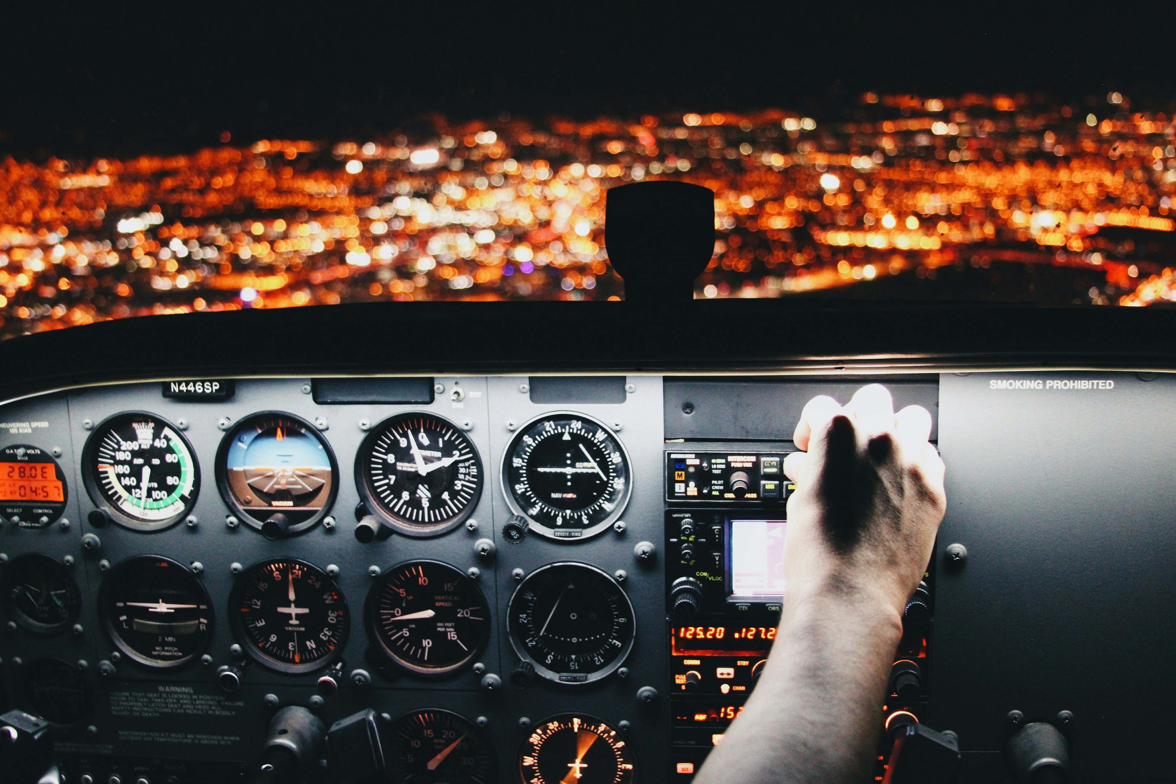 A pilot's hand reaching across the lit instrument panel of a small aircraft cockpit at night, city lights glowing below
